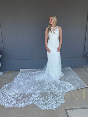 Bride wearing a fitted lace wedding gown with an illusion neckline and intricate floral lace train, standing on a light grey patterned rug against a dark grey wall beside a bridal shop. 