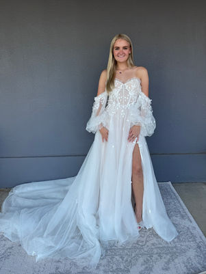 Bride in an ethereal and whimsical off-the-shoulder wedding gown with sheer puff sleeves, a fitted bodice featuring floral appliqués, and a high slit in the skirt, standing on a light grey patterned rug against a dark grey wall beside a bridal shop. 
