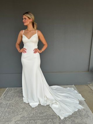 Bride wearing a wedding dress in Shreveport, Bossier City, Minden Louisiana. The wedding dress is a spaghetti strap, fit in flair, Satin sheath dress. Bride standing on a grey patterned rug and grey wall. 