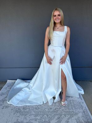 Bride wearing a square-neckline mikado wedding gown, with a fitted bodice and full skirt featuring a high slit, standing on a light grey patterned rug against a dark grey wall beside a bridal shop. 