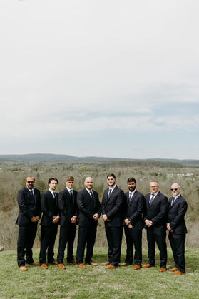 Groom and his groomsmen taking photos after getting married at the ceremony. They are wearing a black tuxedo with a white shirt, black pants, black shoes, Matching vest and bow tie, and a boutonnière. They are standing in a field taking professional wedding photos.