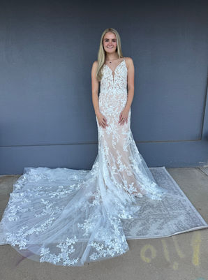 Bride wearing a fitted lace wedding dress with a deep V-neckline, sheer bodice and a long, flowing lace train, standing on a light grey patterned rug against a dark grey wall, beside a bridal shop. 