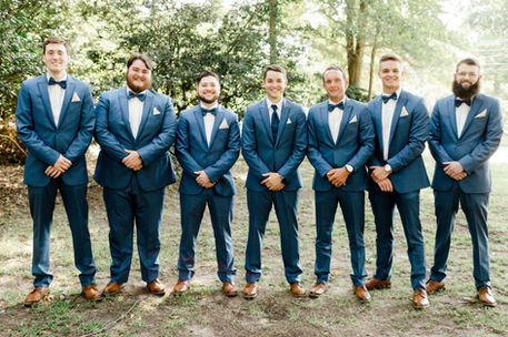 Groom and his groomsmen taking photos after getting married at the ceremony. They are wearing a navy suit with a white shirt, navy pants, brown shoes, Matching vest and bow tie, and a boutonnière. They are standing in a field taking professional wedding photos.