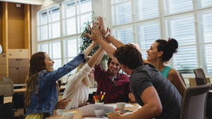 Foto de quatro colegas de trabalho em um escritório, celebrando um momento de sucesso com um 'high five' coletivo sobre a mesa de reunião com papéis e canetas, simbolizando a realização de metas de PDI