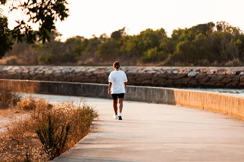 Person in white shirt and black shorts walks away on a curving waterfront path at sunset, with trees and rock wall ahead.