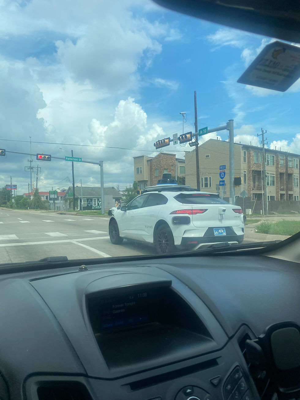 Car interior view of a Waymo white SUV at a stoplight with a blue sky and clouds above. Street sign reads "Patterson St" and "IH 10."