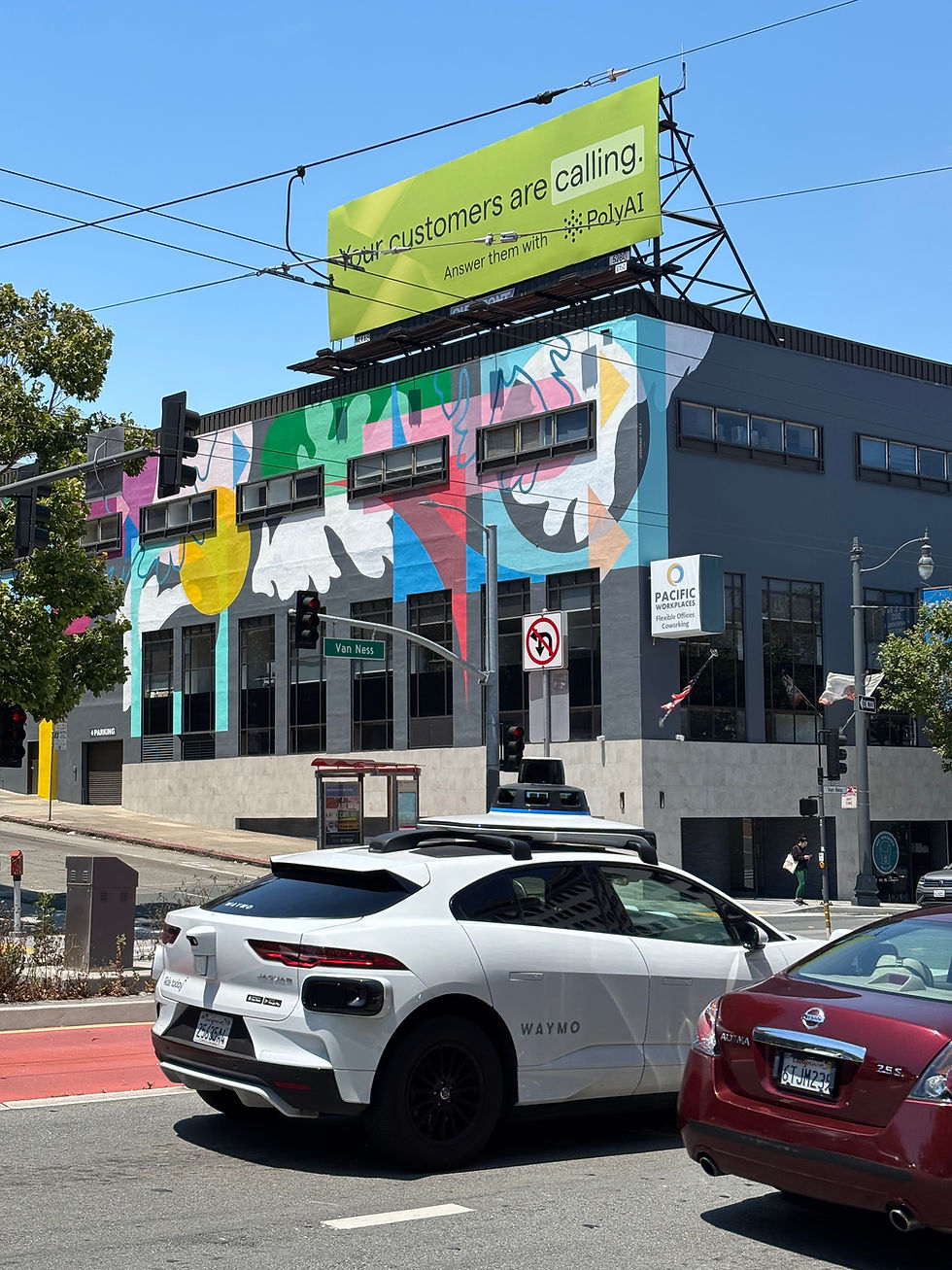 White Waymo car at a colorful mural-covered building with a green billboard reading "Your customers are calling." Clear blue sky above.