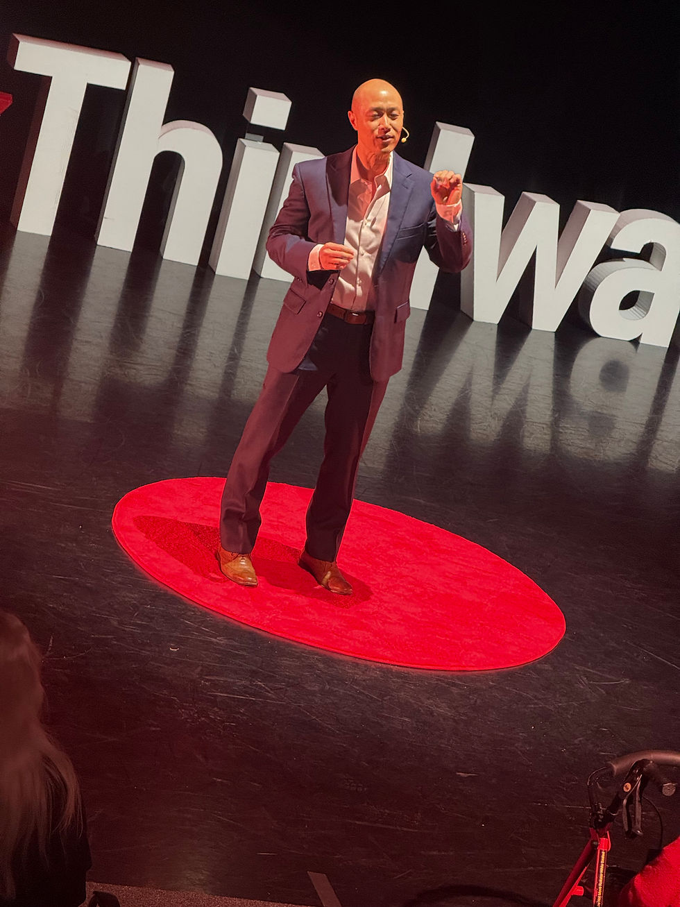 A bald man in a navy suit speaks on a red circle stage, gesturing before large white letters reading Think..., focused and confident.