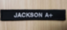 A long, narrow black fabric patch with the name 'JACKSON' and the blood type 'A+' embroidered in white capital letters. The patch is designed to provide identification and critical medical information, set against a light wood grain background.