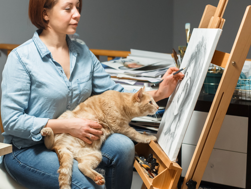 Artist drawing at an easel with a curious orange cat on her lap in a warm, creative studio space.