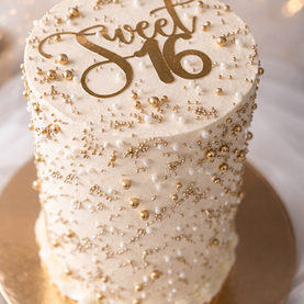 Luxury white buttercream Sweet 16 celebration cake with edible gold and white pearls, textured finish, and gold acrylic topper.