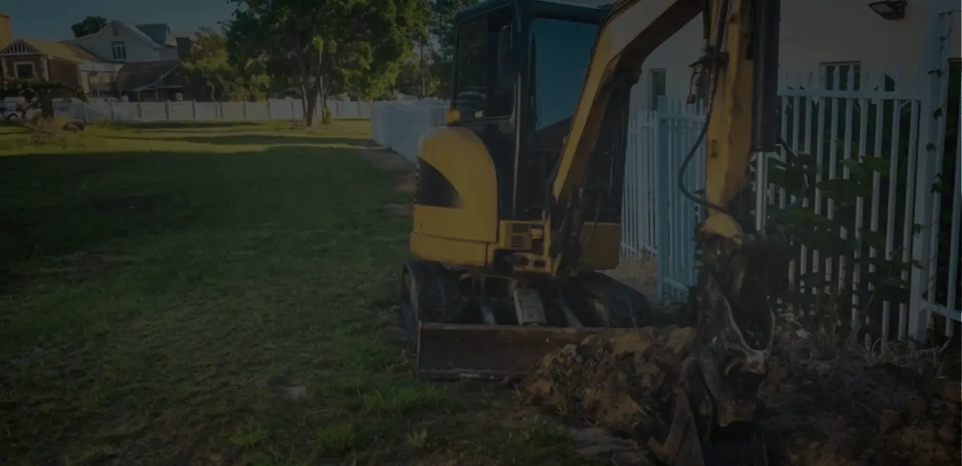 Yellow excavator parked next to a white fence on a residential lawn for site preparation and earthworks on the Gold Coast.