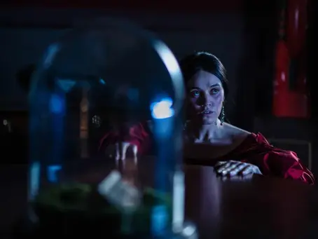 In a dark, blue-lit interior, a woman wearing a red dress sits behind a table, looking intently off-camera. A glass cloche dominates the foreground, creating a sense of mystery and distance.