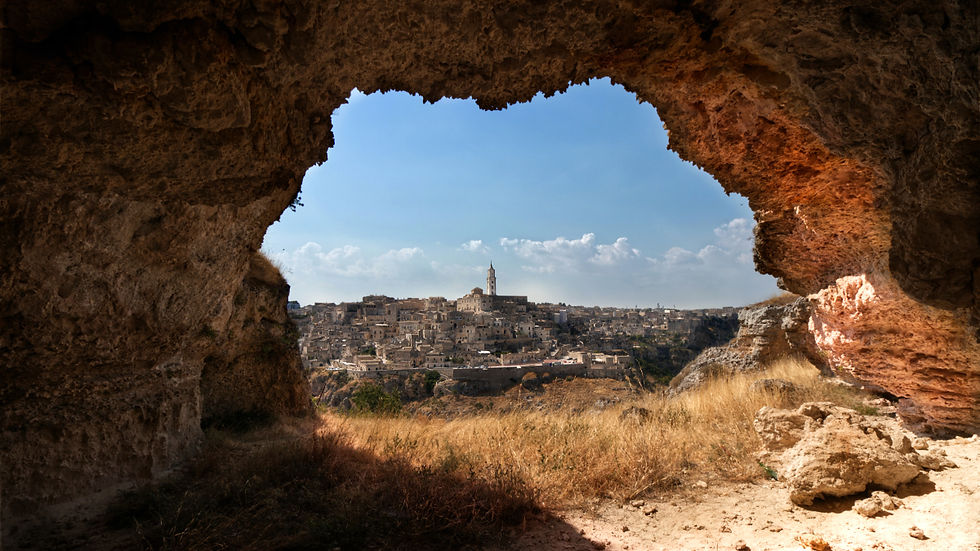 View of an ancient cityscape framed by a cave opening. The sky is clear blue, with a few clouds. Sunlight casts warm tones on the cave walls.