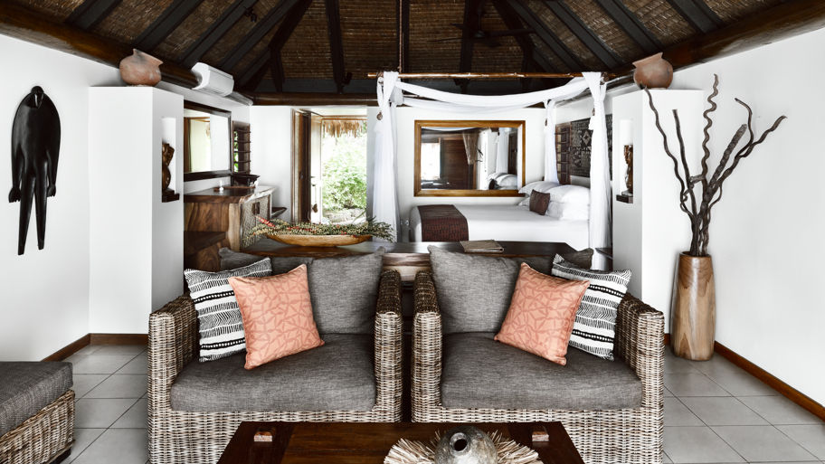 Rustic Fijian Bure living room with wicker sofa and orange pillows, leading to a canopy bed with white drapes. Neutral tones, wooden accents. Cozy vibe.