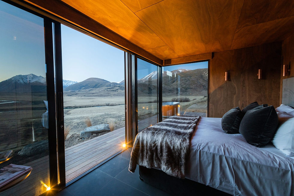 Modern bedroom at The Lindis Lodge with large glass walls overlooking a serene mountain landscape. Warm wood tones inside complement the snowy peaks outside.