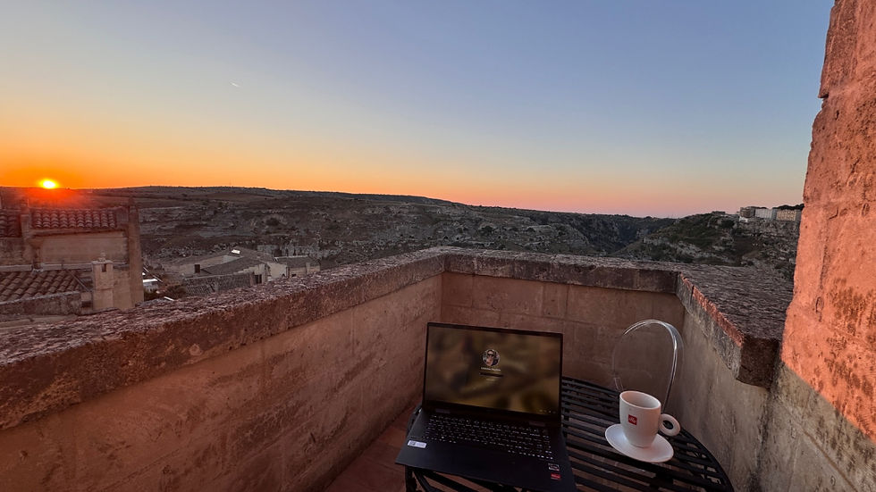 Laptop on a terrace table with a coffee cup at sunset. Overlooks rooftops and a valley under a clear sky with warm, calming hues of Matera.
