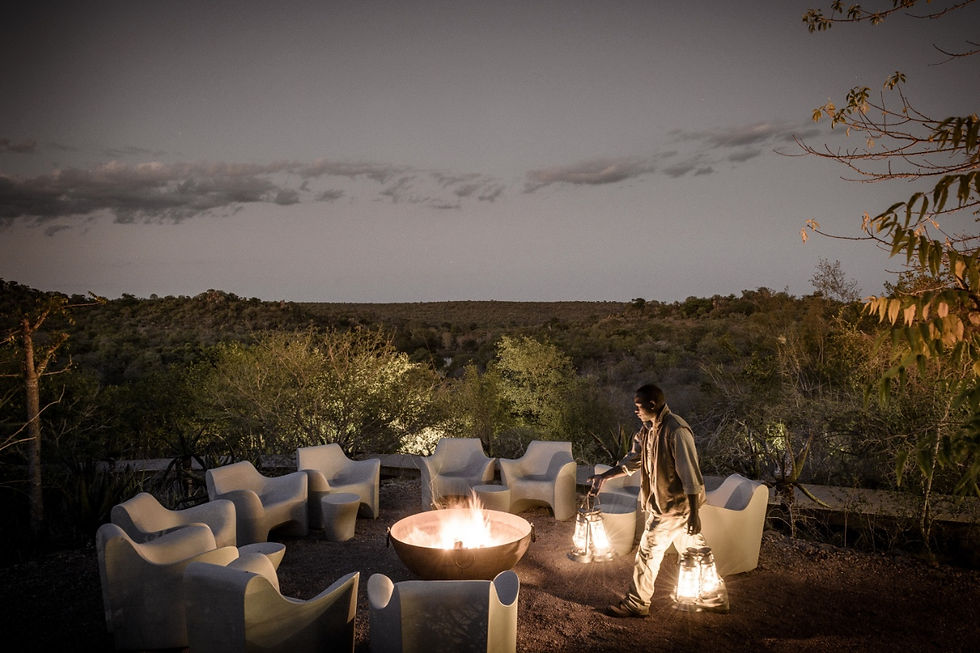 A person carries lanterns near a firepit surrounded by white chairs, set in a dimly lit outdoor area with a scenic landscape backdrop at Singita Lebombo Lodge.