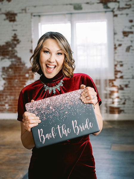 Jenna in a red velvet dress, holding up a laptop with a pink glittery "Back The Bride" written across it.