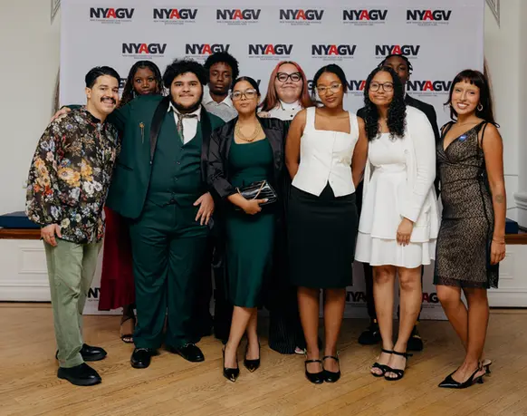 Nine young people in formal attire pose together in front of a step-and-repeat backdrop printed with the NYAGV logo.