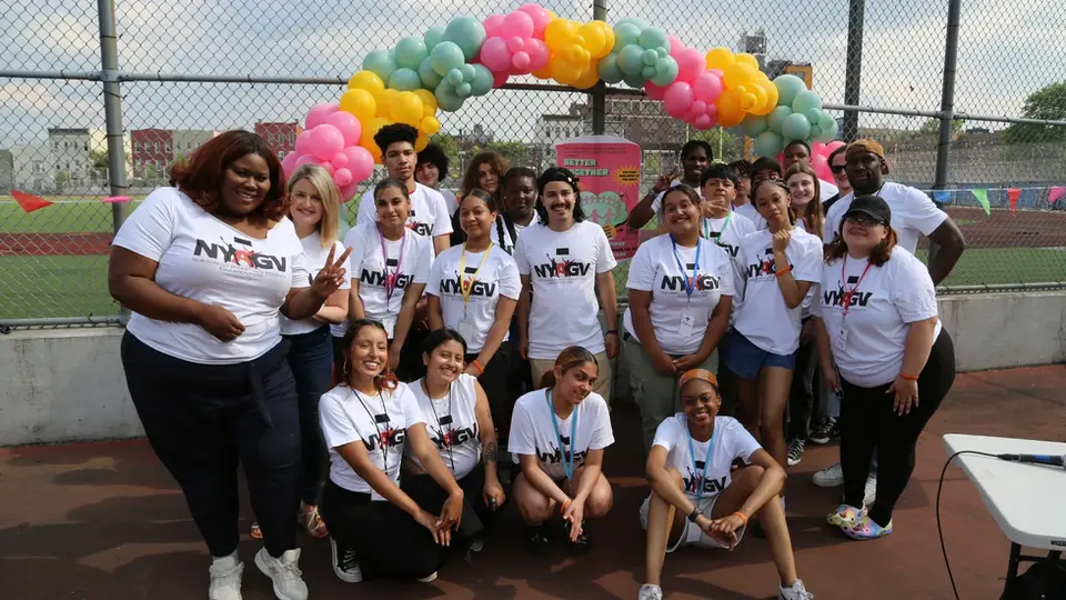 Group of teens and young adults smiling for a photo at a community event, framed by a pink, yellow, and green balloon arch.