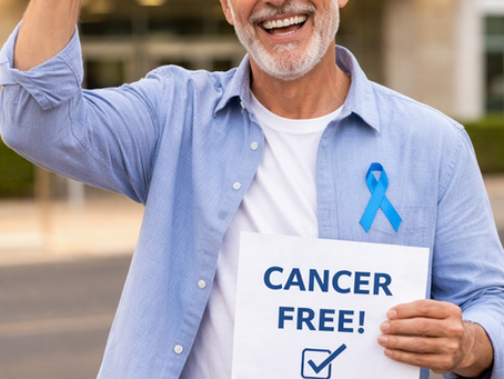 Homem de cerca de 60 anos, cabelos grisalhos e barba branca bem aparada, sorrindo amplamente em frente a um hospital. Ele levanta um dos braços em gesto de vitória e segura um cartaz com a frase “CANCER FREE!” e um símbolo de confirmação. Usa camisa azul clara sobre camiseta branca e um laço azul de conscientização no peito, transmitindo alegria e superação.