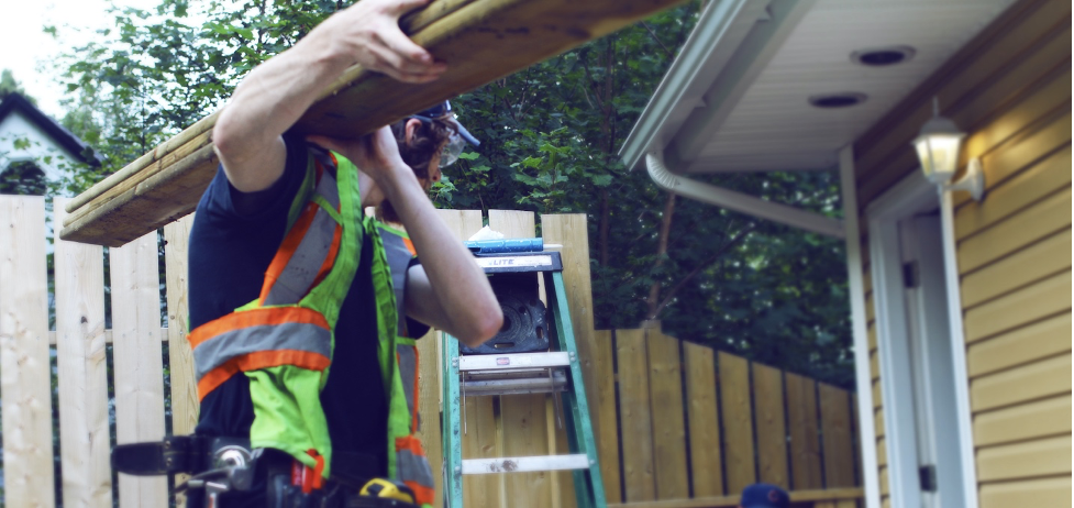 A youth trainee working on a construction site is seen carrying three pieces of lumber towards aYellow house in the background.