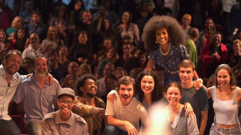 Elenco do Projeto Cria sorrindo no teatro com o público atrás.