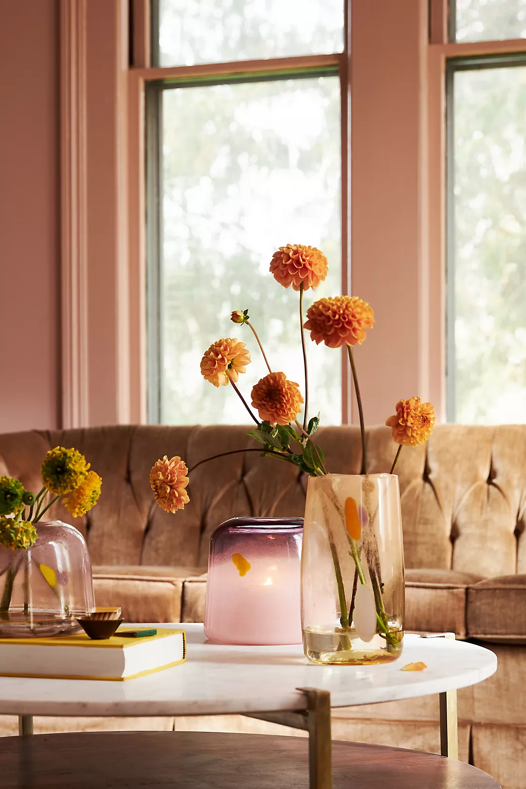 Gezellige interieurhoek met een glazen vaas met oranje bloemen op een salontafel, naast een roze keramische vaas en een elegante bank met tufted bekleding