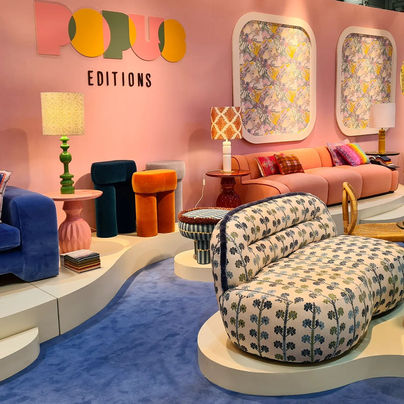 "De afbeelding toont een kleurrijke en stijlvolle interieurshowroom van het merk 'Popus Editions'. De ruimte is ingericht met verschillende meubels, waaronder een blauw fluwelen bank, een oranje fluwelen poef, een beige bank met roze kussens, en een opvallende gestoffeerde chaise longue met een blauw patroon. De achtergrond is een roze muur met drie grote, ingelijste kunstwerken en de merknaam 'Popus Editions' in kleurrijke letters. Er zijn ook verschillende tafellampen met unieke ontwerpen en patronen. De showroom heeft een moderne en speelse uitstraling, met een mix van retro en eigentijdse stijlen.