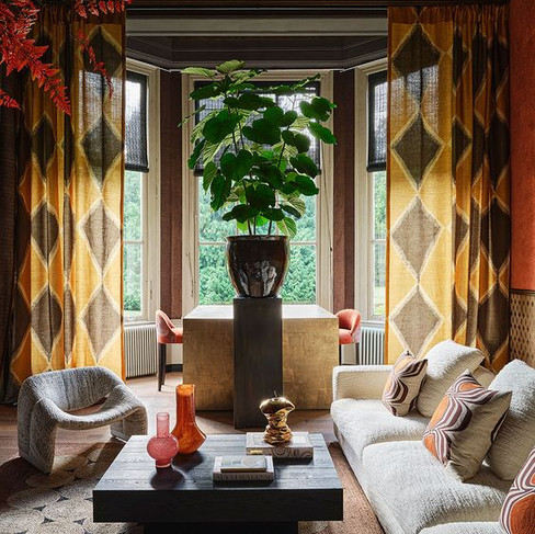 Elegante en gezellige woonkamer met grote ramen, patroon gordijnen en uitzicht op groen gebladerte. De kamer heeft een pluche beige bank met decoratieve kussens, twee unieke grijze fauteuils en een moderne zwarte salontafel met boeken en vazen. De ruimte wordt geaccentueerd door een opvallend beeldhouwwerk op de salontafel, warme verlichting en rijk gekleurde behang, wat een uitnodigende sfeer creëert.