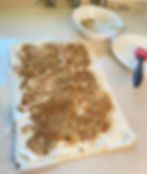 Phyllo dough layered with brown sugar and cinnamon on a beige countertop. White bowl with spoon and plate with red basting brush in background. Warm, baking setup.