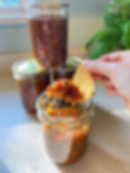 Holding a tortilla chip with a heaping amount of homemade salsa on it hovering above an open jar of salsa and 3 sealed jars glistening in the sunlight pouring in from a window in the background