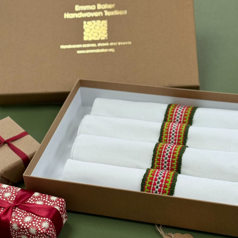 Box of four white napkins with colorful woven bands, set in brown packaging. Nearby are gifts wrapped in patterned and plain paper.