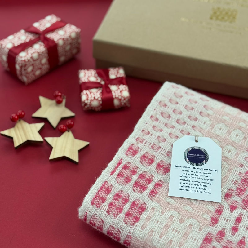 Pink and cream woven textile with Emma Baker tag, surrounded by wrapped gifts and wooden stars on a burgundy background.