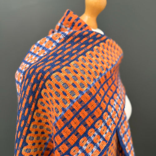 A mannequin displays a vibrant orange and blue patterned scarf with circular and grid designs against a gray background.
