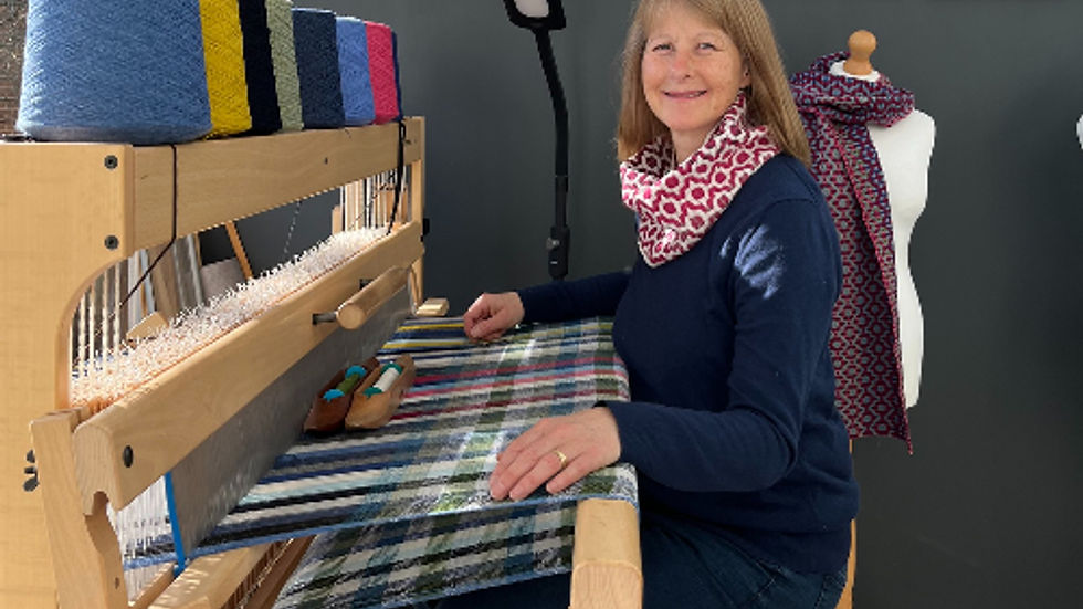 Woman weaving on a loom with colorful threads; wall displays plaid patterns. Bright, cozy room with mannequins; cheerful atmosphere.