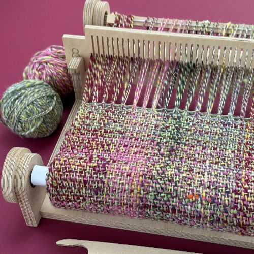 Multicolored woven fabric on a wooden loom with yarn balls beside it against a pink background.