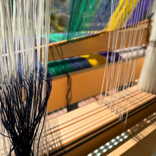 Close-up of a colorful loom with threads in blue, green, yellow, and white. Sunlight casts shadows, creating a serene, artistic mood.