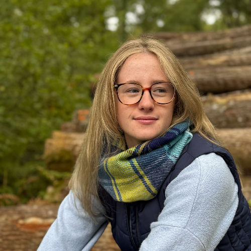 Woman with glasses and plaid scarf sits in front of stacked logs in a forest. She looks thoughtfully to the side. Green and earthy tones.