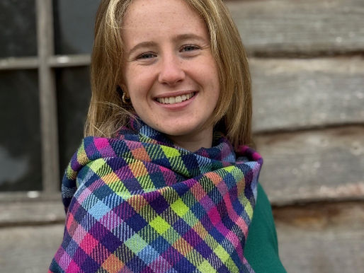 Smiling person wearing a colorful plaid shawl, standing by a wooden wall with a window. Cozy and cheerful atmosphere.