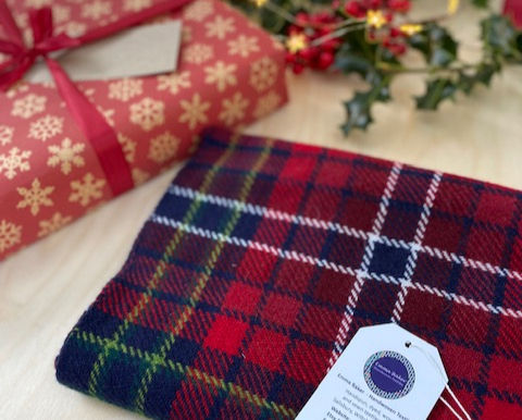 Plaid fabric with a tag next to a red gift box with snowflake pattern. Holly and soft lights in background suggest a festive mood.