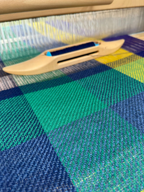 green, blue, purple and yellow scarf being woven on a floor loom with a wooden shuttle