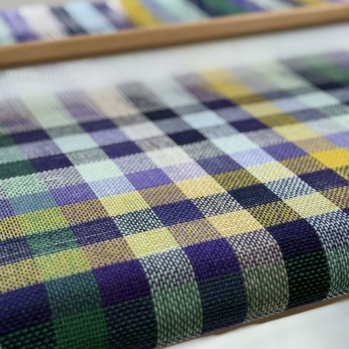 Close-up of a colorful woven fabric on a loom, featuring a checkered pattern in blue, green, yellow, and black hues, creating an artistic vibe.