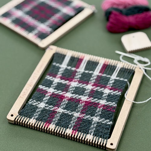 Plaid woven fabric on a small wooden loom with pink and black yarn, on a green table, with yarn balls and tools nearby