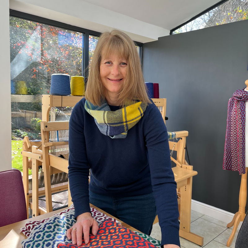 Woman smiling in a bright room with looms and colorful yarn. She wears a yellow plaid scarf and touches patterned fabric on a table.