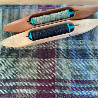 Weaving on the floor loom in green and burgundy shades with two weaving shuttles at the top of the picture.