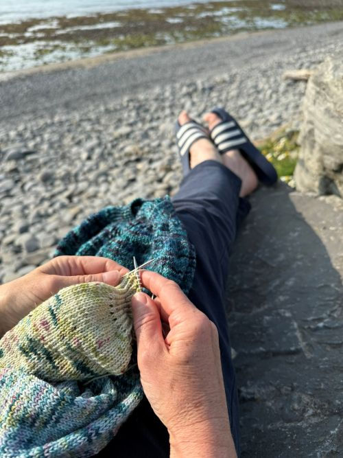 Hands knitting a colorful pattern by the rocky shore, with relaxed legs and sandals visible. Calm seaside setting with a serene mood.