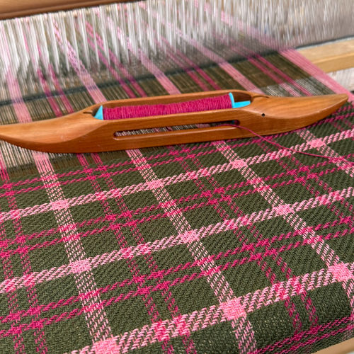 Wooden weaving shuttle with pink yarn on a loom, over green-and-pink plaid fabric in progress.
