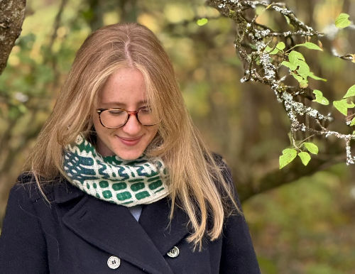 Woman with glasses in a navy coat and green scarf, smiling softly, stands in a leafy forest with a serene mood.